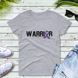 Puede incluir: Una camiseta gris jaspeado con la palabra "WARRIOR" en letras negras y un lazo morado. La camiseta se muestra sobre una superficie de madera clara, con otros objetos visibles.