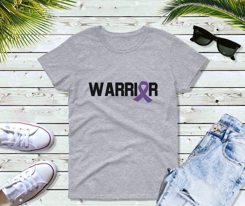 Puede incluir: Una camiseta gris jaspeado con la palabra "WARRIOR" en letras negras y un lazo morado. La camiseta se muestra sobre una superficie de madera clara, con otros objetos visibles.