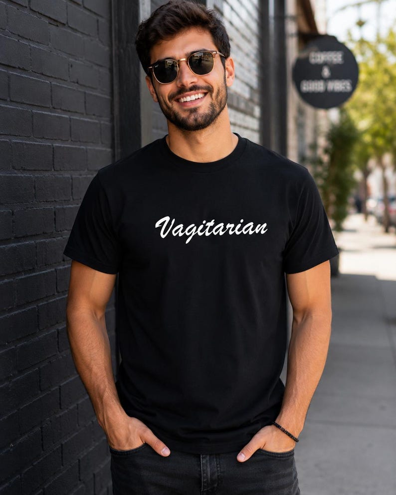 May include: Black t-shirt with the word "Vagitarian" in white script. The shirt is made of a soft material. The person in the image is wearing sunglasses and has a beard. The background includes a black brick wall and a sign that says "Coffee & Good Vibes."