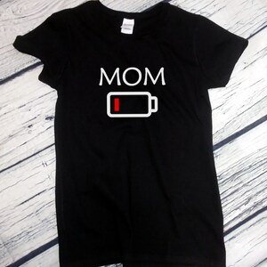 Pu&ograve; includere: T-shirt nera con la scritta "MOM" in lettere maiuscole bianche. Sotto, un simbolo di batteria bianco con una barra rossa che indica la batteria scarica. La maglietta &egrave; esposta su una superficie in legno chiaro.