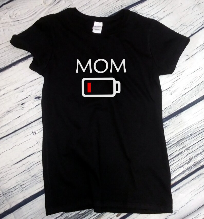 Pu&ograve; includere: T-shirt nera con la scritta "MOM" in lettere maiuscole bianche. Sotto, un simbolo di batteria bianco con una barra rossa che indica la batteria scarica. La maglietta &egrave; esposta su una superficie in legno chiaro.