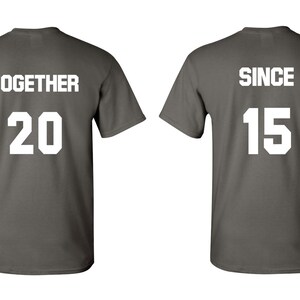 Op de afbeelding: Twee grijze T-shirts met witte tekst. Het eerste shirt zegt "TOGETHER 20" en het tweede shirt zegt "SINCE 15".