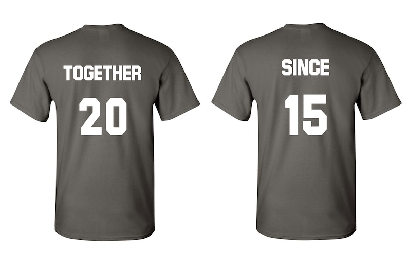 Op de afbeelding: Twee grijze T-shirts met witte tekst. Het eerste shirt zegt "TOGETHER 20" en het tweede shirt zegt "SINCE 15".