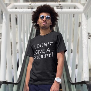 May include: A black t-shirt with white text that reads "I don't give a schnitzel". The person wearing the shirt is standing on an escalator and wearing sunglasses.