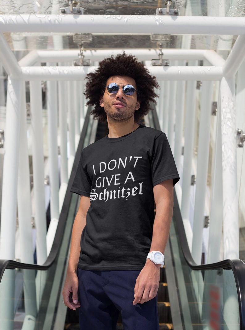 May include: A black t-shirt with white text that reads "I don't give a schnitzel". The person wearing the shirt is standing on an escalator and wearing sunglasses.