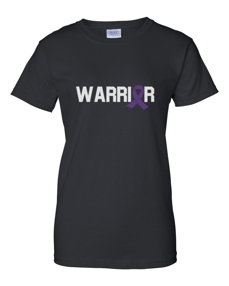 Puede incluir: Camiseta negra con la palabra "WARRIOR" en letras blancas y una cinta morada de concienciaci&oacute;n. La camiseta es de cuello redondo y est&aacute; hecha de un material suave. Corte cl&aacute;sico.