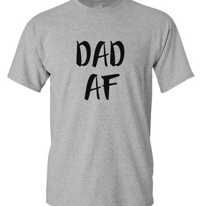 Peut inclure: Un t-shirt gris chin&eacute; avec l'inscription "DAD AF" en lettres noires grasses. Le t-shirt a des manches courtes et un col rond.