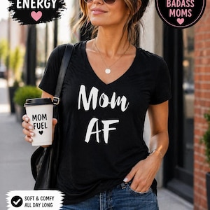 May include: Black V-neck t-shirt with the words "Mom AF" in white script. The image includes text that says "Bold Mom Energy" and "The perfect gift for badass moms". The shirt is paired with blue jeans.