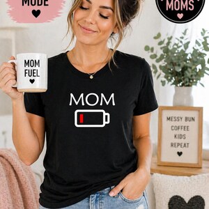 Pu&ograve; includere: Maglietta nera con grafica bianca "MOM" e simbolo di batteria scarica. Viene tenuta una tazza bianca con "MOM FUEL". L'immagine include il testo "Mom Life Mode" e "Perfect Gift for Moms".