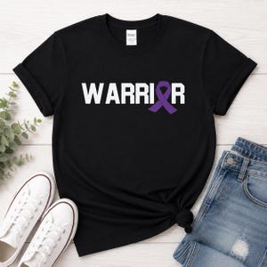 Puede incluir: Camiseta negra con la palabra "WARRIOR" en letras blancas y una cinta morada. La camiseta se combina con vaqueros azules y zapatillas de lona blancas. Las mangas est&aacute;n dobladas.