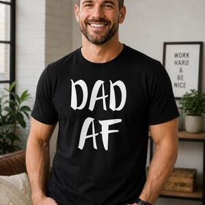 Peut inclure: T-shirt noir avec le texte blanc "DAD AF". Le t-shirt est port&eacute; par une personne. Un panneau encadr&eacute; avec le texte "WORK HARD & BE NICE" est visible en arri&egrave;re-plan.