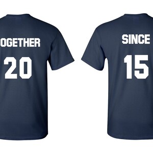 Op de afbeelding: Twee donkerblauwe T-shirts met witte tekst. Het eerste shirt zegt "TOGETHER 20" en het tweede shirt zegt "SINCE 15".