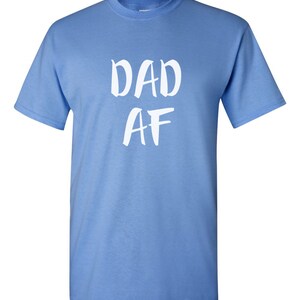 Peut inclure: Un t-shirt bleu clair avec le texte blanc "DAD AF" &eacute;crit &agrave; la main. Le t-shirt a des manches courtes et un col rond.