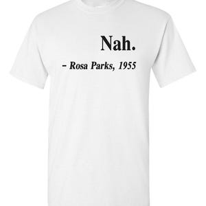 May include: White t-shirt with the word "Nah" in a bold, black font at the top center. Below the word is the text "- Rosa Parks, 1955" in a smaller, black font. The shirt is a classic crew neck style.