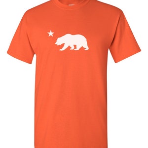 May include: Orange short-sleeved t-shirt with a white silhouette of a bear and a star.
