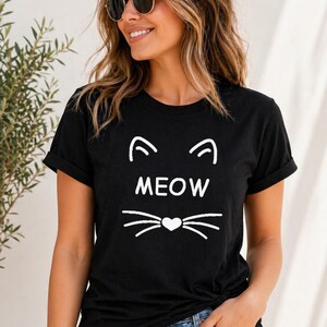 Puede incluir: Camiseta negra con un dise&ntilde;o de cara de gato blanco. El dise&ntilde;o incluye orejas de gato, la palabra "MEOW" y bigotes con un coraz&oacute;n. La camiseta tiene mangas cortas y cuello redondo.
