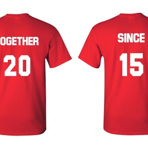 Op de afbeelding: Twee rode T-shirts met witte tekst. Het eerste shirt zegt "TOGETHER 20" en het tweede shirt zegt "SINCE 15".