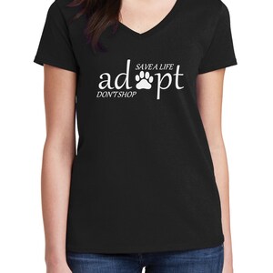 May include: A black V-neck t-shirt featuring white text that reads "adopt" with a paw print replacing the letter "o", accompanied by the smaller text "SAVE A LIFE" above the paw and "DON'T SHOP" positioned below the word "adopt".