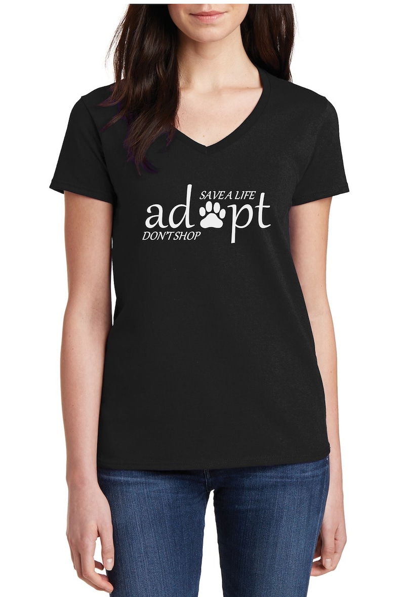 May include: A black V-neck t-shirt featuring white text that reads "adopt" with a paw print replacing the letter "o", accompanied by the smaller text "SAVE A LIFE" above the paw and "DON'T SHOP" positioned below the word "adopt".