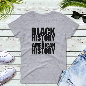 May include: A heather gray t-shirt with the text "BLACK HISTORY IS AMERICAN HISTORY" in bold, black capital letters. The shirt is displayed on a light wood-grain surface, with white sneakers, sunglasses, and blue jeans.