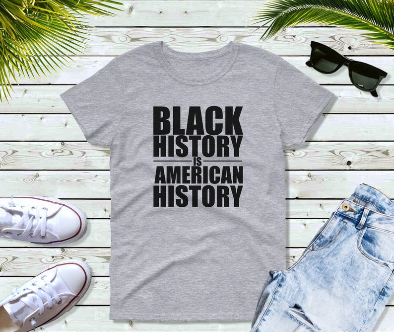 May include: A heather gray t-shirt with the text "BLACK HISTORY IS AMERICAN HISTORY" in bold, black capital letters. The shirt is displayed on a light wood-grain surface, with white sneakers, sunglasses, and blue jeans.