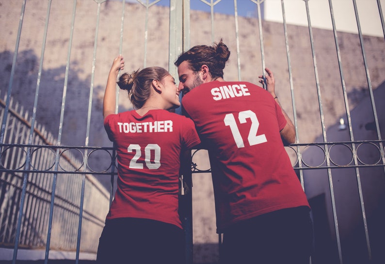 Op de afbeelding: Een stel draagt ​​bijpassende rode T-shirts met witte tekst. Het shirt van de vrouw zegt "Together 20" en het shirt van de man zegt "Since 12". Ze staan ​​voor een metalen hek.