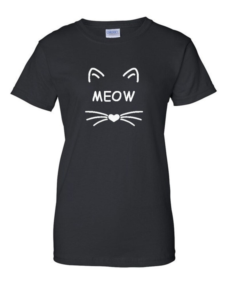 Puede incluir: Camiseta negra con un dise&ntilde;o de cara de gato blanco. El dise&ntilde;o incluye dos l&iacute;neas curvas para las orejas, la palabra "MEOW" y bigotes con una nariz en forma de coraz&oacute;n.