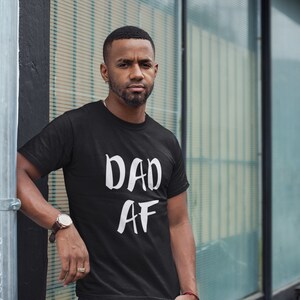Peut inclure: T-shirt noir avec le texte blanc "DAD AF". Le t-shirt &agrave; manches courtes semble &ecirc;tre en coton. La personne portant le t-shirt est &agrave; l'ext&eacute;rieur, pr&egrave;s d'un poteau m&eacute;tallique et d'une fen&ecirc;tre en verre.