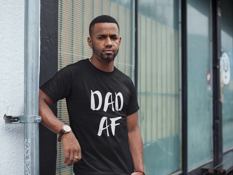 Peut inclure: T-shirt noir avec le texte blanc "DAD AF". Le t-shirt &agrave; manches courtes semble &ecirc;tre en coton. La personne portant le t-shirt est &agrave; l'ext&eacute;rieur, pr&egrave;s d'un poteau m&eacute;tallique et d'une fen&ecirc;tre en verre.