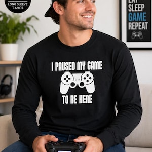 May include: Black long-sleeve t-shirt with the text "I paused my game to be here" and a white controller graphic. The shirt also has the text "Eat Sleep Game Repeat" on a framed print. The shirt is a Gildan 5400 long sleeve t-shirt.