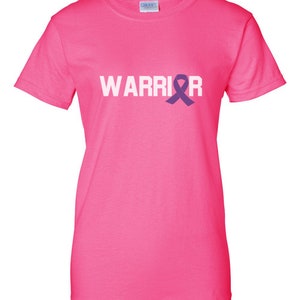 Puede incluir: Una camiseta rosa brillante con la palabra "WARRIOR" impresa en blanco y un lazo morado. La camiseta tiene cuello redondo y est&aacute; hecha de un tejido suave.