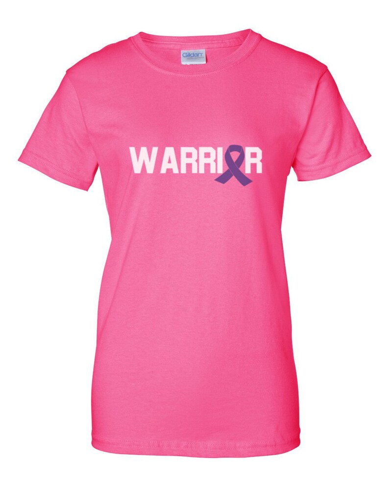 Puede incluir: Una camiseta rosa brillante con la palabra "WARRIOR" impresa en blanco y un lazo morado. La camiseta tiene cuello redondo y est&aacute; hecha de un tejido suave.