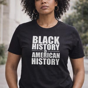 May include: Black t-shirt with the text "BLACK HISTORY IS AMERICAN HISTORY" in white block letters. The shirt is worn by a person with curly dark hair and gold hoop earrings.