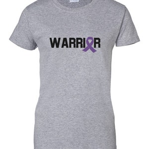 Puede incluir: Una camiseta gris jaspeado con la palabra "WARRIOR" impresa en negro y una cinta morada de concienciaci&oacute;n. La camiseta es de cuello redondo y est&aacute; hecha de un material suave. Es una prenda informal.