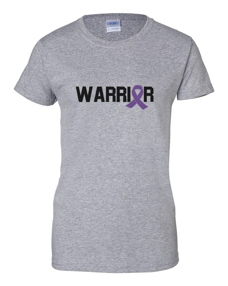 Puede incluir: Una camiseta gris jaspeado con la palabra "WARRIOR" impresa en negro y una cinta morada de concienciaci&oacute;n. La camiseta es de cuello redondo y est&aacute; hecha de un material suave. Es una prenda informal.