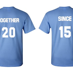 Op de afbeelding: Twee lichtblauwe T-shirts met witte tekst. Het linker T-shirt zegt "TOGETHER 20" en het rechter T-shirt zegt "SINCE 15".
