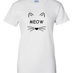 Puede incluir: Camiseta blanca con un dise&ntilde;o de cara de gato negro. El dise&ntilde;o incluye l&iacute;neas curvas para las orejas, la palabra "MEOW" en letras negritas y bigotes con una nariz en forma de coraz&oacute;n. La camiseta es de material suave.