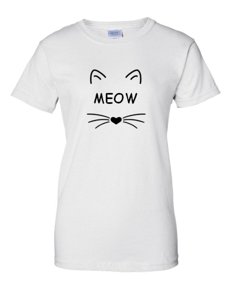 Puede incluir: Camiseta blanca con un dise&ntilde;o de cara de gato negro. El dise&ntilde;o incluye l&iacute;neas curvas para las orejas, la palabra "MEOW" en letras negritas y bigotes con una nariz en forma de coraz&oacute;n. La camiseta es de material suave.