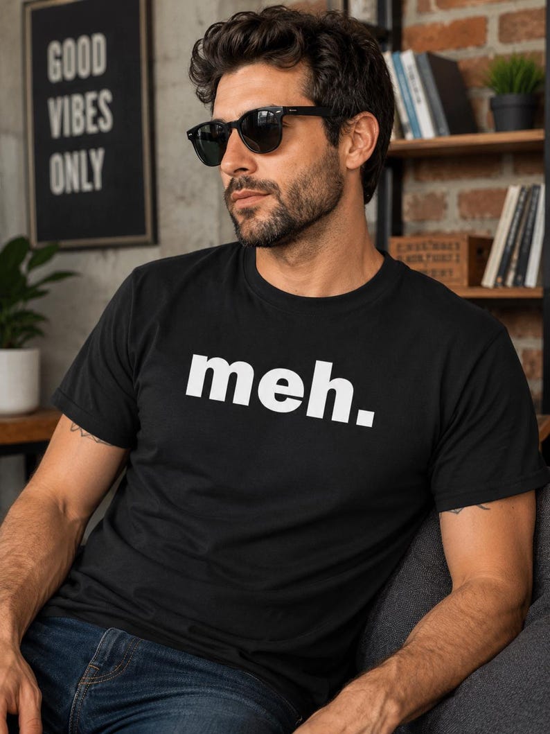 May include: Black t-shirt with the word "meh." in white block letters. The person is wearing black sunglasses and blue jeans. A sign in the background reads "GOOD VIBES ONLY."