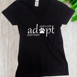 May include: A black V-neck t-shirt featuring white text that reads "adopt" with a paw print replacing the letter "o", "SAVE A LIFE" above the paw, and "DON'T SHOP" below the word "adopt". The shirt has short sleeves and a Gildan brand label.