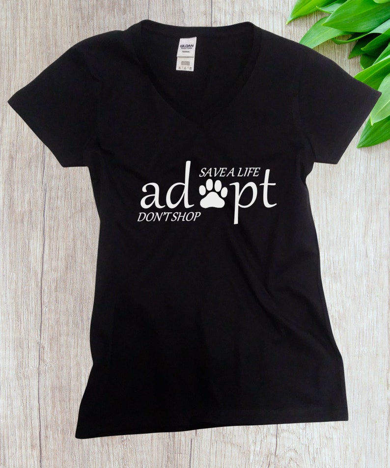 May include: A black V-neck t-shirt featuring white text that reads "adopt" with a paw print replacing the letter "o", "SAVE A LIFE" above the paw, and "DON'T SHOP" below the word "adopt". The shirt has short sleeves and a Gildan brand label.