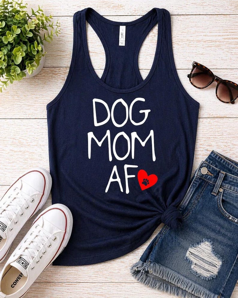 May include: Navy blue tank top with the words "DOG MOM AF" in white, a red heart with a paw print, and a racerback design. The image also includes white sneakers, denim shorts, and sunglasses.