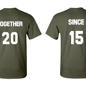 Op de afbeelding: Twee olijfgroene T-shirts met witte tekst. Het eerste shirt zegt "TOGETHER 20" en het tweede shirt zegt "SINCE 15".