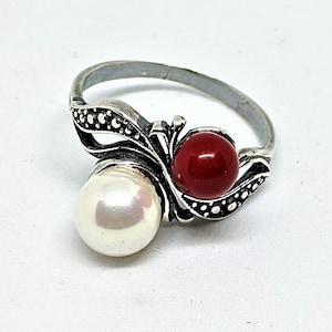 May include: A silver ring with a white pearl and a red stone. The ring has a delicate design with a floral motif.