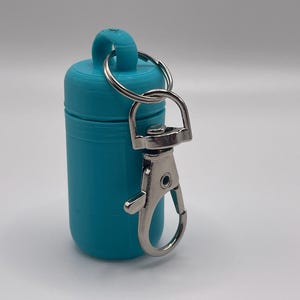 May include: A teal, cylindrical container with a loop on top, attached to a silver-colored metal clip and key ring. The container appears to be made of plastic. The clip is open.