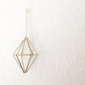 May include: A gold geometric ornament hanging from a white ceiling. The ornament is shaped like a diamond and is made of thin metal rods.