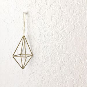 May include: A gold geometric hanging ornament with a diamond shape. The ornament is made of thin metal rods and hangs from a chain.