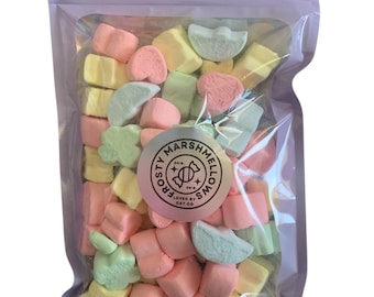 May include: A clear plastic bag filled with pastel-colored marshmallows in the shapes of hearts, stars, and half-moons. The bag has a purple zipper and a pink and white label that reads "FROSTY MARSHMALLOWS" with a pink and white candy cane graphic.