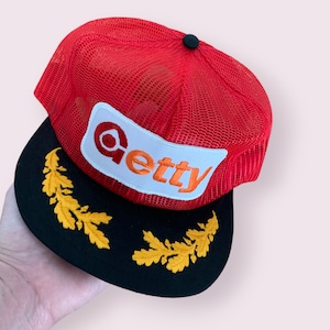 May include: Red mesh trucker cap with a black peak. The cap has a white patch with the name "Getty" in orange and red lettering. The peak has embroidered gold leaf designs.