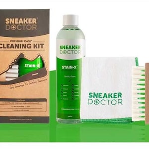 Puede incluir: Un kit de limpieza para zapatillas con una etiqueta verde y blanca que dice "Sneaker Doctor" y una botella de limpiador con una etiqueta verde que dice "Stain-X". El kit también incluye un paño de microfibra blanco y un cepillo.